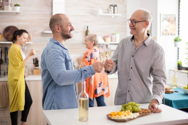 Felice figlio e padre che tintinnano bicchieri di vino in cucina durante il pranzo con la famiglia.