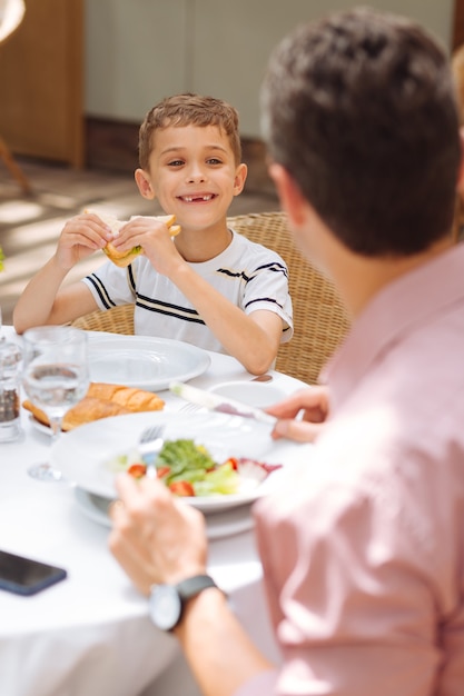 Figlio felice. piccolo figlio felice che mangia delizioso panino al pollo durante la colazione in famiglia fuori casa