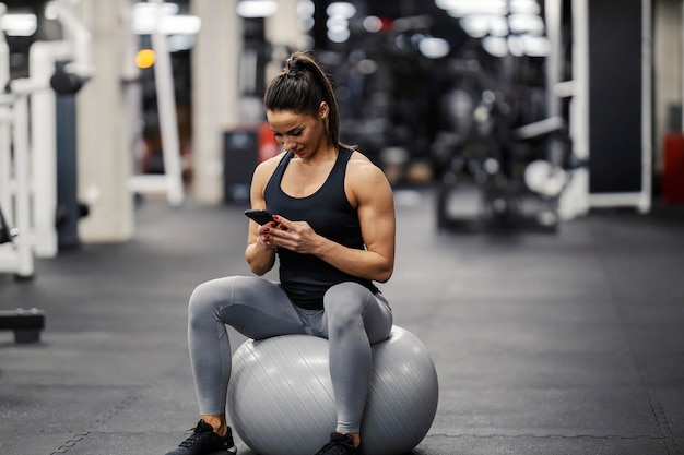 A happy sportswoman is sitting on a fitness ball in a gym and checking out new fitness app