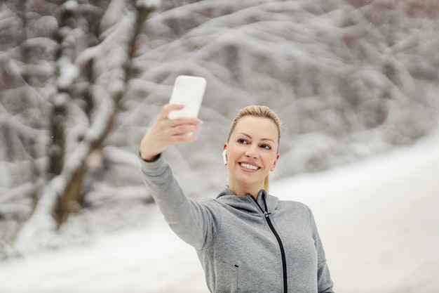 雪の降る冬の日に自然の中で立って、ソーシャルメディアのために自分撮りをしている幸せなスポーツウーマン。