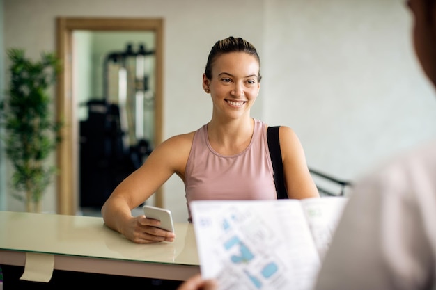 Happy sportswoman using smart phone while talking to receptionist at health club