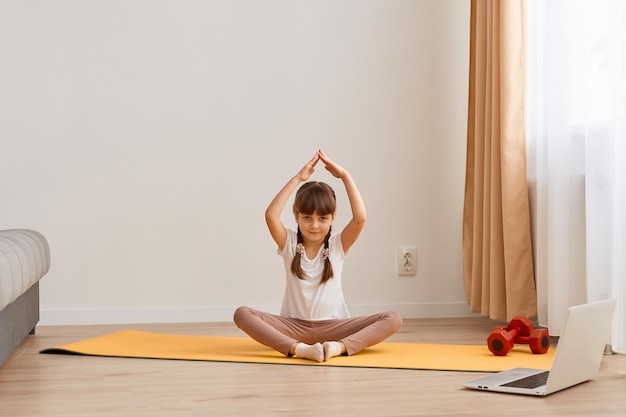 Happy sporty Caucasian little girl meditating in living room healthy child with raised arms sitting on the floor yoga mat in lotus position looking at camera with kind positive expression