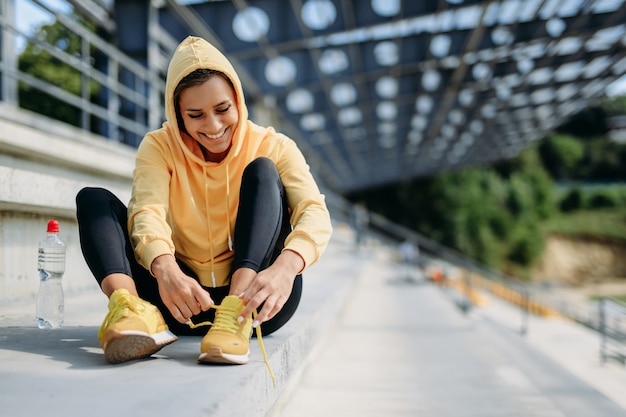 Happy sporty female in yellow hoodie and black leggings sitting on stairs and tying laces on sneakers.