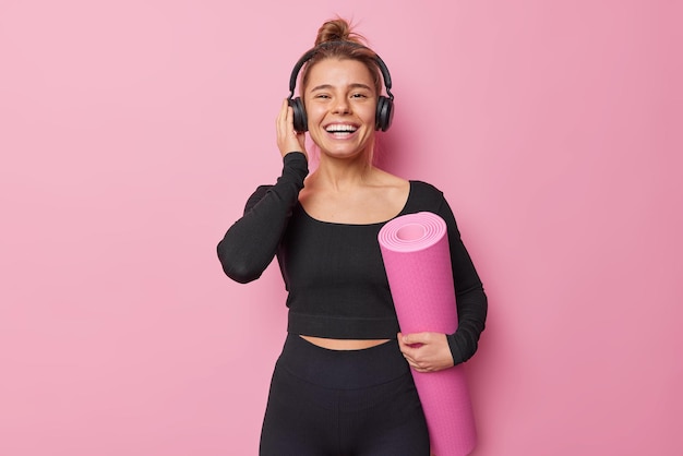 Happy sporty woman dressed in black sportswear carries rolled karemat poses at yoga studio going to have practice enjoys good sound in headphones isolated over pink background. Fitness concept