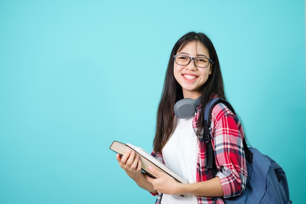 幸せな学生。青い背景の上のスタジオでバックパックと立っているカメラに微笑んで陽気なアジアの女の子。学校のコンセプトに戻ります。