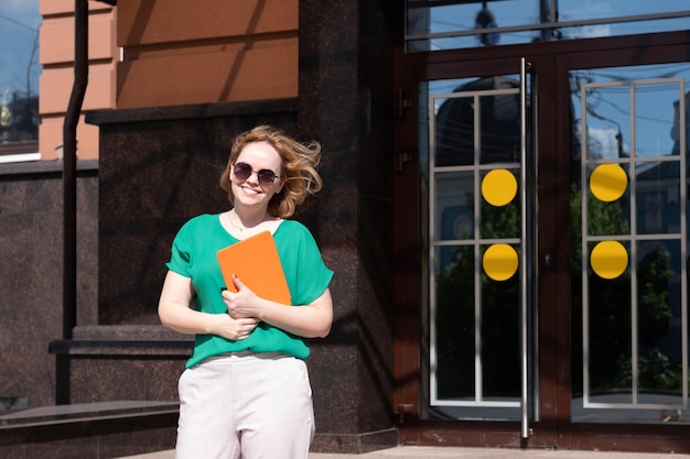 A happy student girl in sunglasses holding a digital tablet book noutbook against the university building