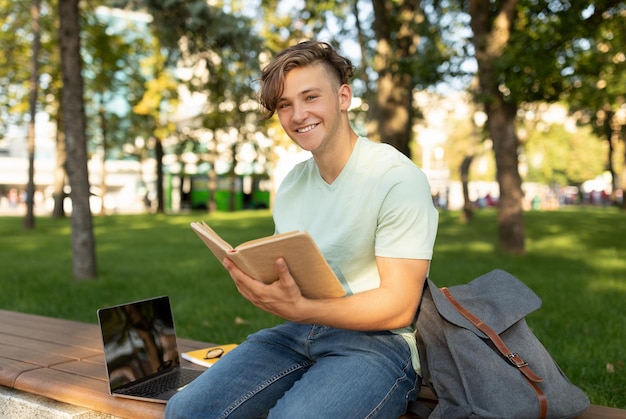 Happy student guy reading interesting book having rest after classes sitting on bench in parkland on