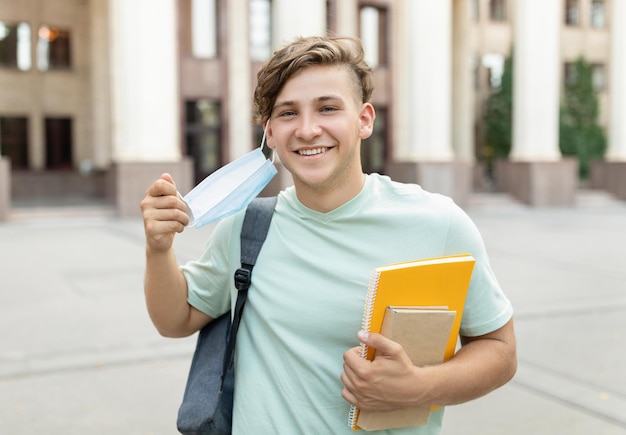 Happy student guy removing medical mask posing outdoors with backpack and books standing near