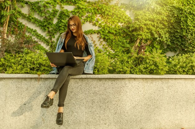 Happy student woman in denim jacket and glasses uses laptop while sitting in the city park