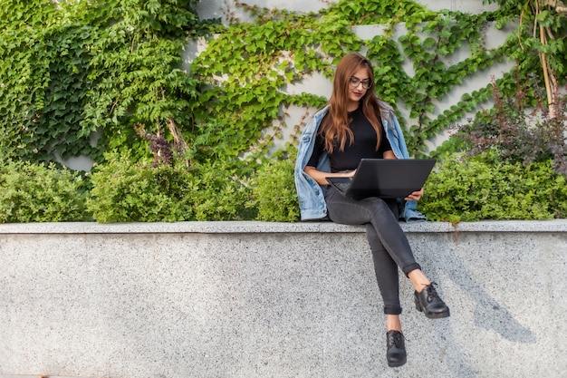 Счастливый студент женщина в джинсовой куртке и очках использует ноутбук, сидя в городском парке