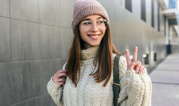 Donna felice dello studente sorridente che indossa un maglione e occhiali trasparenti alla moda che mostrano un gesto di pace bella giovane donna felice che posa fuori alla luce del sole nella strada della città