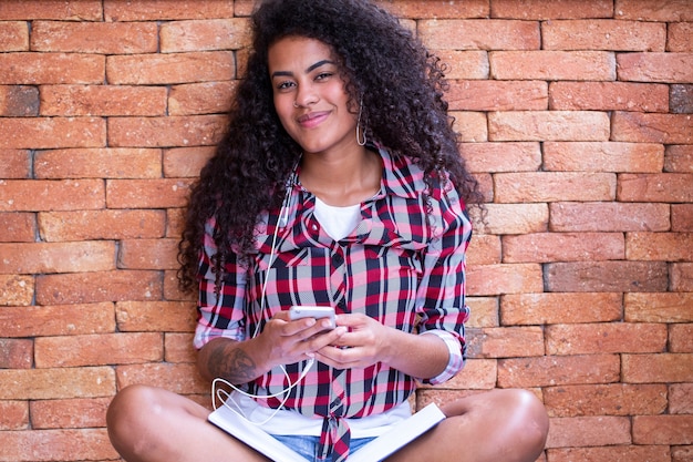 Happy student woman with afro hairstyle sitting with brick wall background using cell phone and smiling.