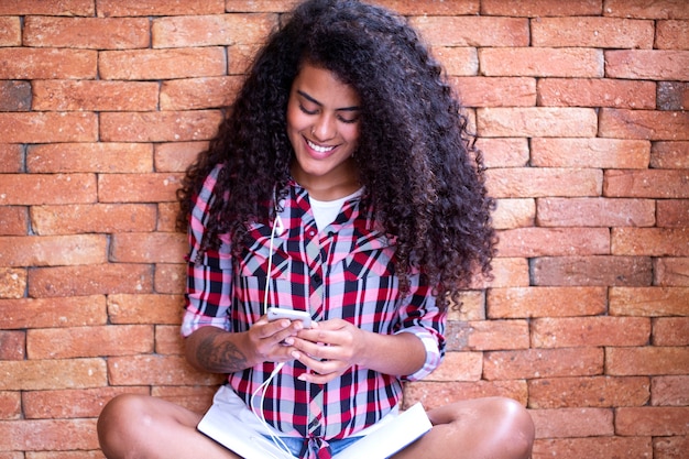 Foto felice studente donna con acconciatura afro seduto con sfondo muro di mattoni utilizzando il telefono cellulare e sorridente.