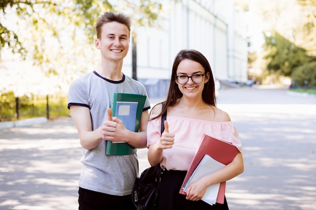 Coppia di studenti felici in piedi in un parco e mostrando i pollici in su