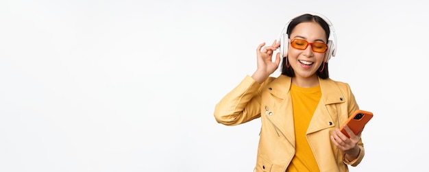 Happy stylish asian woman in sunglasses listening music in headphones holding smartphone singing and