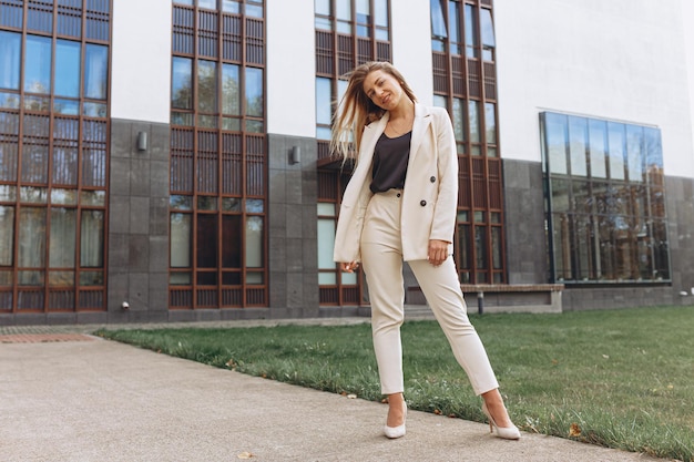 Happy stylish businesswoman wearing white elegant suit and high heels standing against building