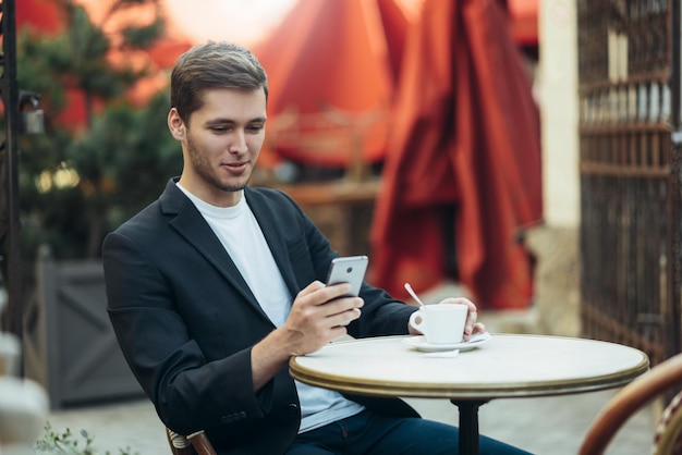 Happy stylish Caucasian businessman in suit drinking cappccino at coffee shop sitting on terrace with cell phone Business modern technology communication and people concept