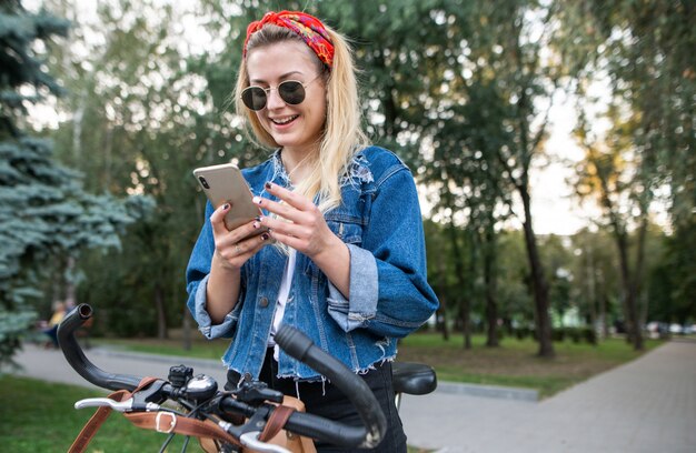 公園で自転車に座って、スマートフォンを使用して、笑顔の幸せでスタイリッシュな女の子。公園の自転車の上を歩きます。