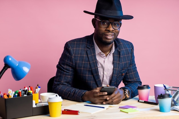 Happy stylish handsome afro american businessman sitting at desk surrounded with stationery, wearing glasses, suit, black hat, holding smartphone, sending email, reading news, over pink background