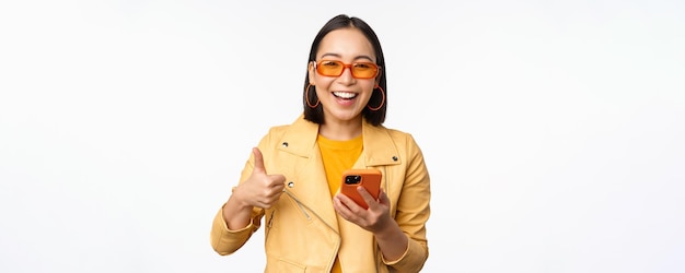 Happy stylish korean girl wearing sunglasses showing thumb up holding smartphone using mobile phone