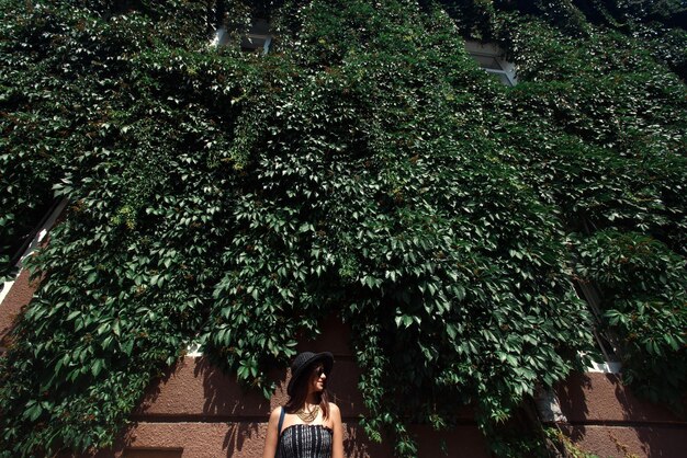 写真 日当たりの良い街の夏の旅行の概念の緑の葉の壁の建物に立っている幸せなスタイリッシュな女性ヒップスター