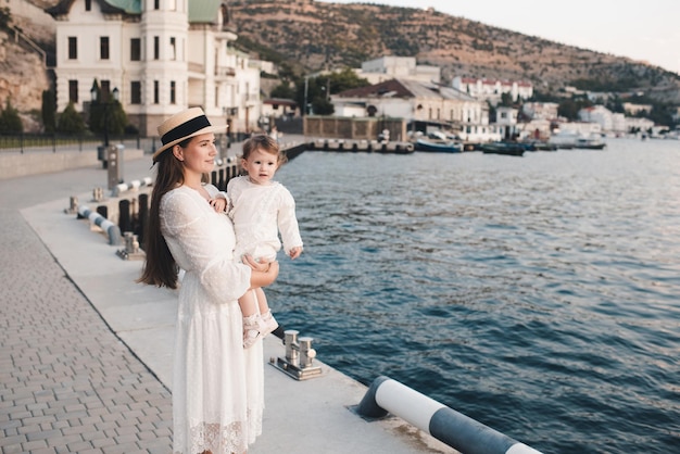 Happy stylish woman hold baby girl 1 year old wear white trendy dress and straw hat walk on quayside over sea shore outdoors together. Motherhood. Maternity. Summer vacation time.