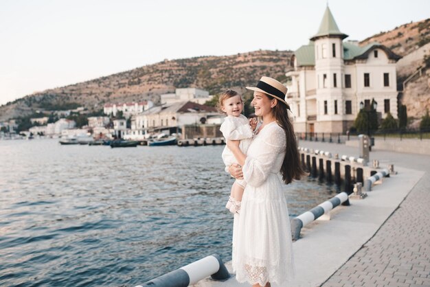 Happy stylish woman hold baby girl 1 year old wear white trendy dress and straw hat walk on quayside over sea shore outdoors together. Motherhood. Maternity. Summer vacation time.