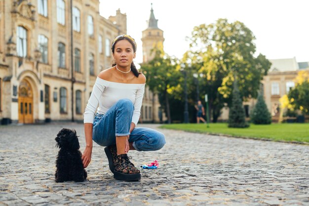 カメラを見ている小さなトイプードル犬の後ろの舗装に座っている幸せなスタイリッシュな女性