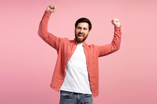 Happy stylish young man in shirt celebrating with arms up looking at camera isolated on pink