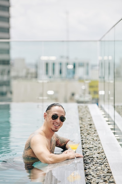 Счастливый стильный молодой человек в солнцезащитных очках, проводящий время в бассейне на крыше с бокалом коктейля