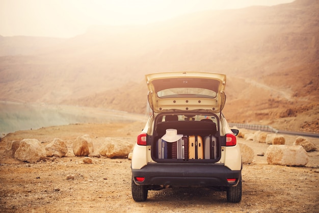 Happy stylish young woman traveler on beach road sitting on white crossover car