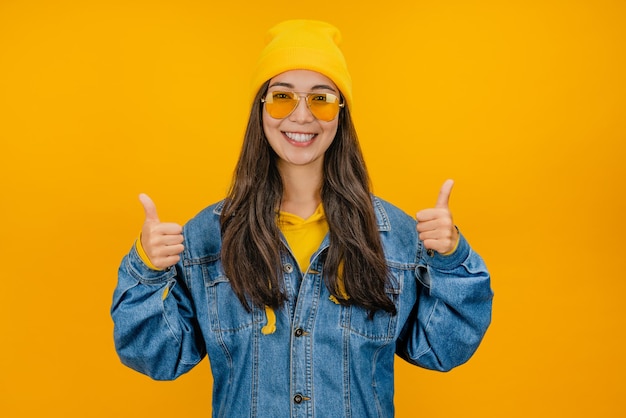 Happy stylish young woman in yellow hat and sunglasses showing thumbs up on yellow background