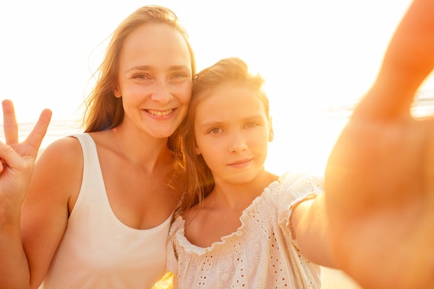 Счастливые стильные мать и дочь делают селфи на песчаном пляже на закате. Маленькая блондинка и красивая женщина фотографируют на телефон.