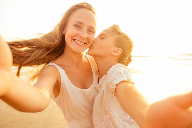 幸せなスタイリッシュな母のキス娘と日没の砂浜で自分撮りを撮る。母の日。小さな女の子のブロンドと美しい女性が海外の電話観光オンラインビデオ通話で写真を撮る。