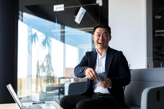 Счастливый и успешный азиатский бизнесмен, работающий в офисе с ноутбуком, счастливый, держа наличные доллары, рвет, празднуя победу и победу