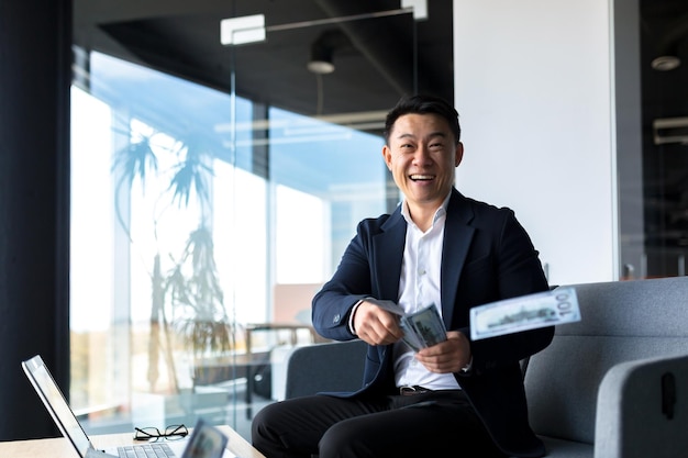 Счастливый и успешный азиатский бизнесмен, работающий в офисе с ноутбуком, счастливый, держа наличные доллары, рвет, празднуя победу и победу