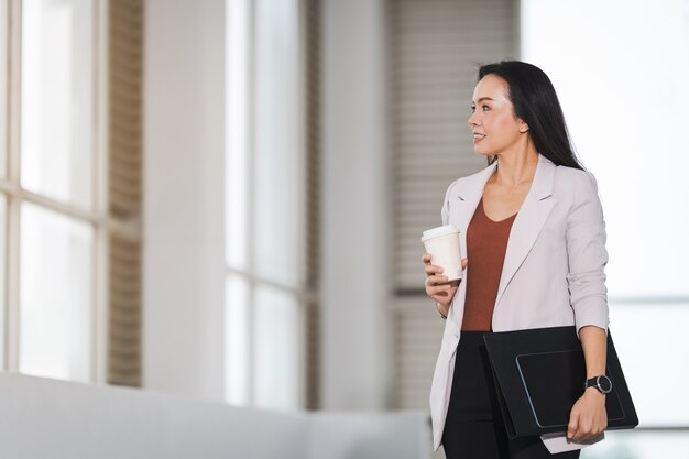 A happy successful Asian businesswoman holds a takeaway coffee cup and document in business building