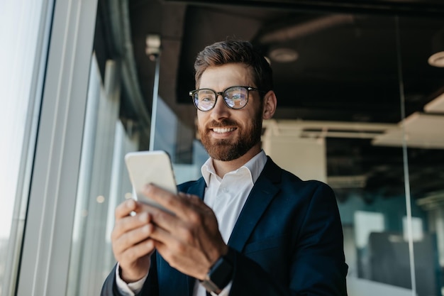 Happy successful businessman in glasses typing on smartphone in modern office interior having chat with client online