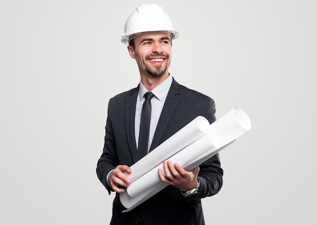 Happy successful male construction architect in elegant suit and hardhat with rolled drafts in hands looking away and smiling against white background