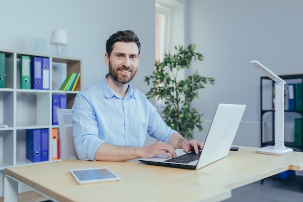 Happy and successful man working on laptop in home office sitting at table in bright room smiling looking at camera