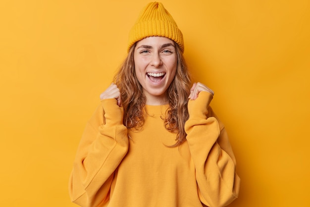 Happy successful woman clenches fists celebrates triumph smiles pleased laughs joyfully wears hat and casual jumper isolated over vivid yellow background shouts with joyful face yes i did it