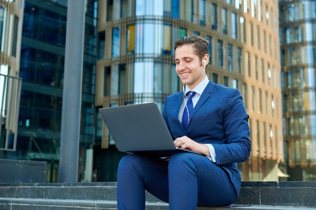 Happy successful young businessman working on modern laptop, using earphones and sitting on stairs outside skyscrapers