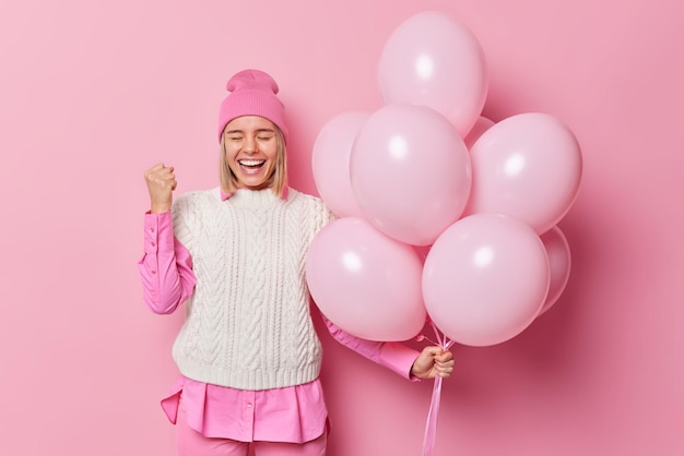 Happy successful young woman celebrates success or special occasion wears hat and vest comes on party holds bunch of inflated helium balloons isolated over pink background makes winner gesture