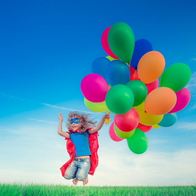 Happy superhero child jumping with colorful toy balloons outdoors. Smiling kid having fun in green spring field against blue sky background. Freedom concept