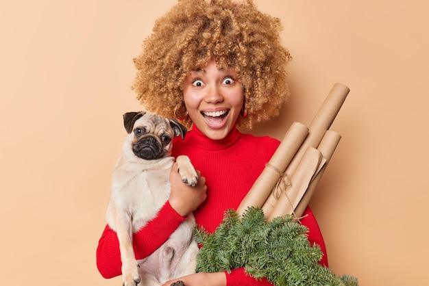 La donna felice e sorpresa sente una grande notizia tiene le pose del cane carlino con carta decorativa e la corona di abete verde sembra sbalordita isolata su sfondo beige. reazioni umane emozioni preparazione per le vacanze