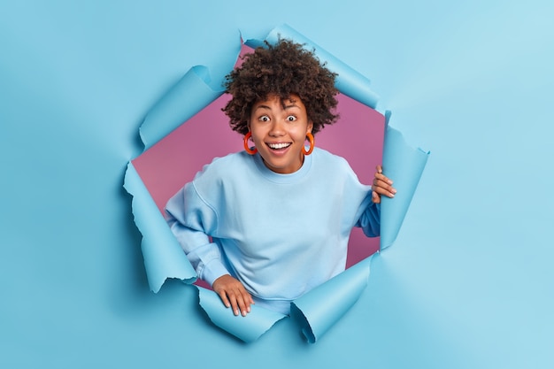 Happy surprised Afro American teenage girl reacts on something awesome smiles broadly poses in torn paper blue wall dressed in casual jumper