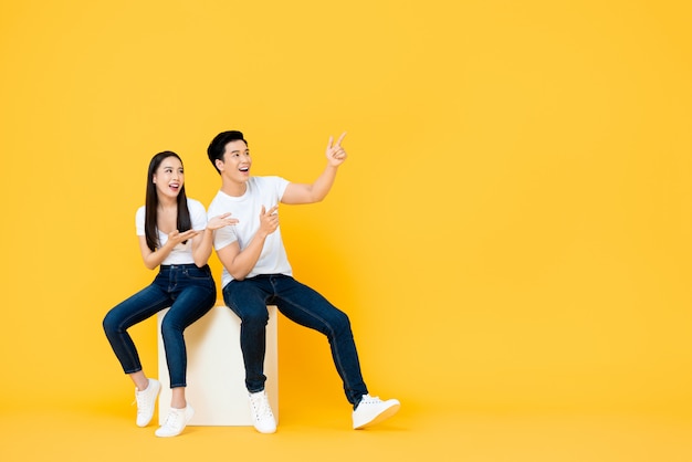 Happy surprised attractive young Asian couple pointing and looking at blank space beside in yellow isolated stuidio wall