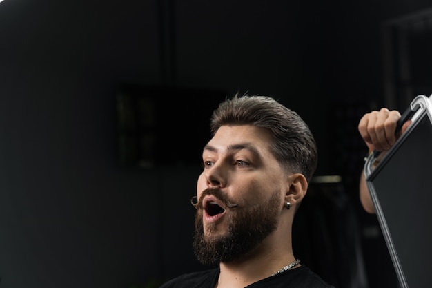 Happy and surprised bearded man looking in mirror in barbershop. Showing result of low fade machine hair.