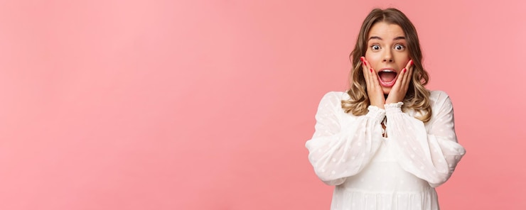 Photo happy and surprised blond girl hear awesome news gasping speechless staring camera with hands on cheeks being impressed and stunned over great event standing pink background