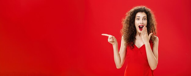 Foto felice sorpresa e deliziata femmina dai capelli ricci attraente in abito da sera sorridente stupito con ope